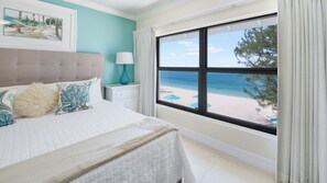 Un dormitorio con un ventanal amplio que ofrece vista a la playa, una cama con ropa blanca y una mesita de noche con lámpara.
