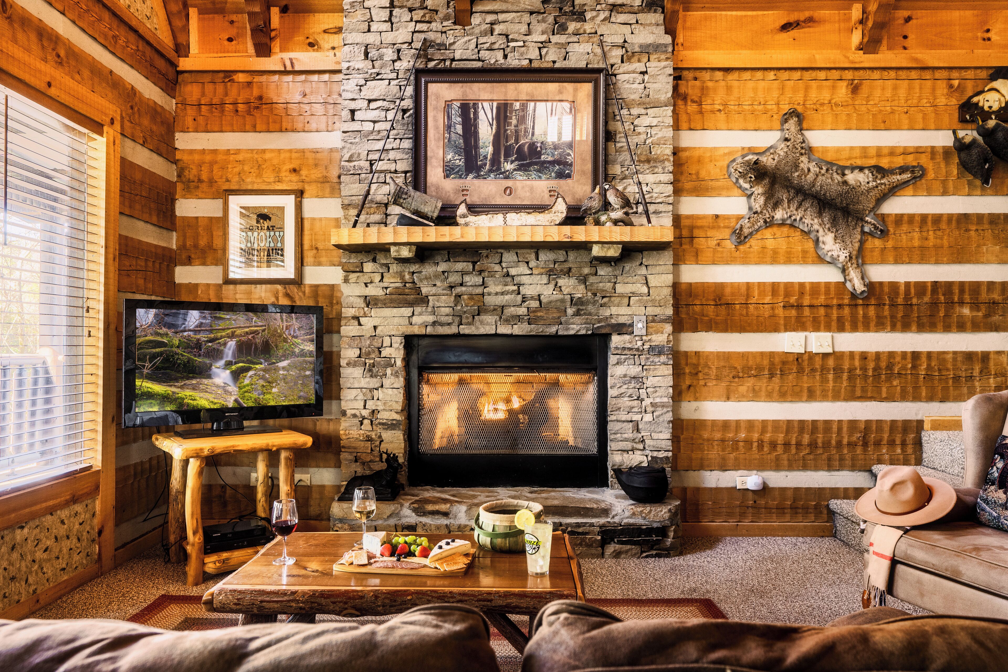 A cozy log cabin interior with a stone fireplace, a television, and a dining area with a table set for two.