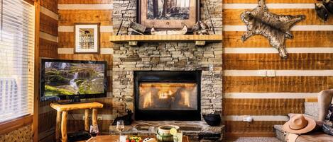 A cozy log cabin interior with a stone fireplace, a television, and a dining area with a table set for two.