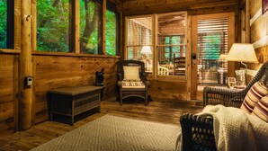A cozy room with wooden walls, a wicker chair, a bench, and a view of greenery through large windows. - Up The Creek Cabin - Peaceful surroundings on a mountain stream (Sevierville)
