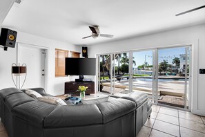 A modern living room with a large sectional sofa, a flat-screen TV, ceiling fans, and a view of a pool and palm trees. - Coquina Paradise: waterfront, pool, hot tub, boat dock and lift (St. Petersburg)