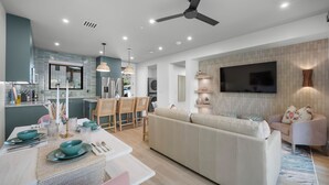 Una sala moderna con un sofá, un comedor con una mesa preparada para dos, y una cocina con refrigerador y lavadora.