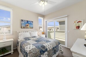 A bedroom with a bed, a ceiling fan, a window with blinds, a painting, and a view through a sliding door.