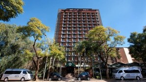 Un hotel alto con fachada uniforme, múltiples balcones y un estacionamiento con varios vehículos.