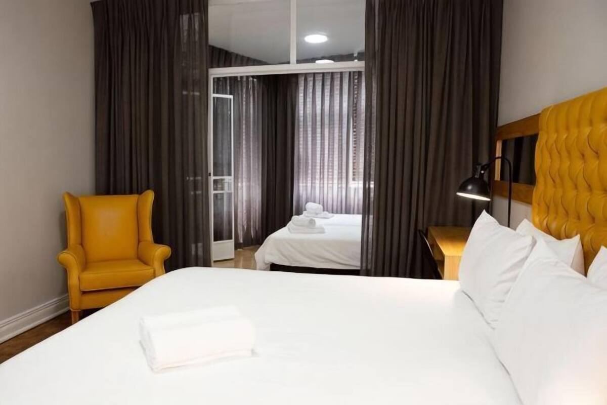 Habitación de hotel con una cama grande, un sillón amarillo, una mesita de noche con lámpara y vistas a través de un ventanal.