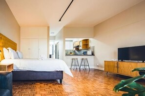 A bedroom with a bed, a TV, a wooden floor, and a kitchen area in the background. - Hotel 224 & Apartments (Pretoria)