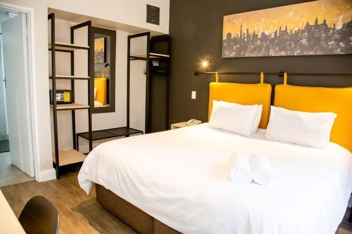 Una habitación de hotel con una cama, un cuadro de paisaje urbano, una estantería y una silla.