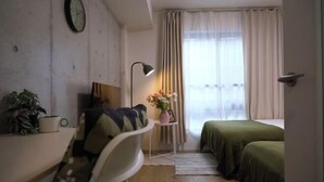 A small, cozy room with a bed, a desk, a chair, a lamp, a clock, and a window with curtains. - Poka Stay Kamata (Tokyo)