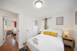 Quarto com cama, mesas de cabeceira, janela com cortinas e porta que dá para outro cômodo.