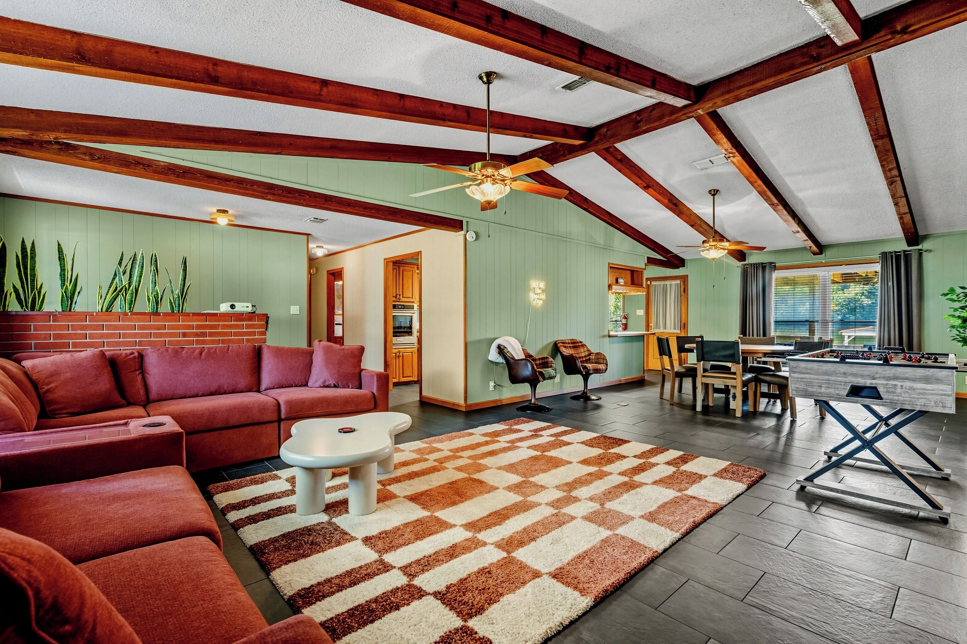 A spacious living area with a red sofa, a coffee table, and a checkered rug. There is a foosball table and a dining area with chairs and a table.