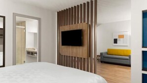 Ett modernt hotellrum med en stor säng, en väggmonterad TV, en väggpanel i trä, en grå soffa med en gul kudde och en inramad tavla på väggen.