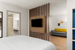 A hotel room with a bed, a flat-screen TV mounted on a wooden panel, a gray sofa with a yellow cushion, and a wooden floor. - Holiday Inn Express SAN JOSE FORUM by IHG (Pozos)