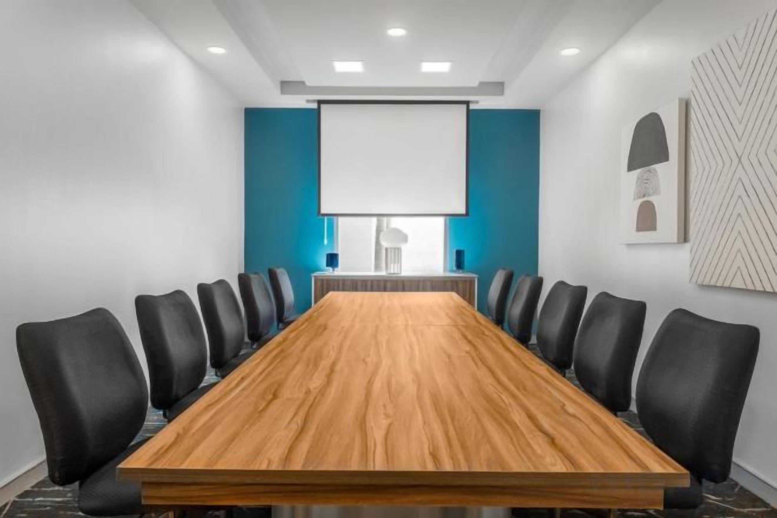 Un salón de conferencias con una larga mesa, sillas negras, una pantalla de proyector y una pared azul de acento.