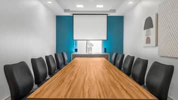 Un salón de conferencias con una larga mesa, sillas negras, una pantalla de proyector y una pared azul de acento.