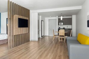 A modern living room with a flat-screen TV mounted on a wooden panel, a grey sofa with a yellow cushion, and a wooden dining table with chairs. - Holiday Inn Express SAN JOSE FORUM by IHG (Pozos)