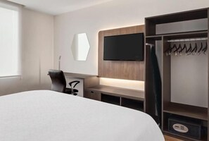 A hotel room with a bed, a desk, a chair, a TV mounted on the wall, and a wardrobe with hanging clothes.