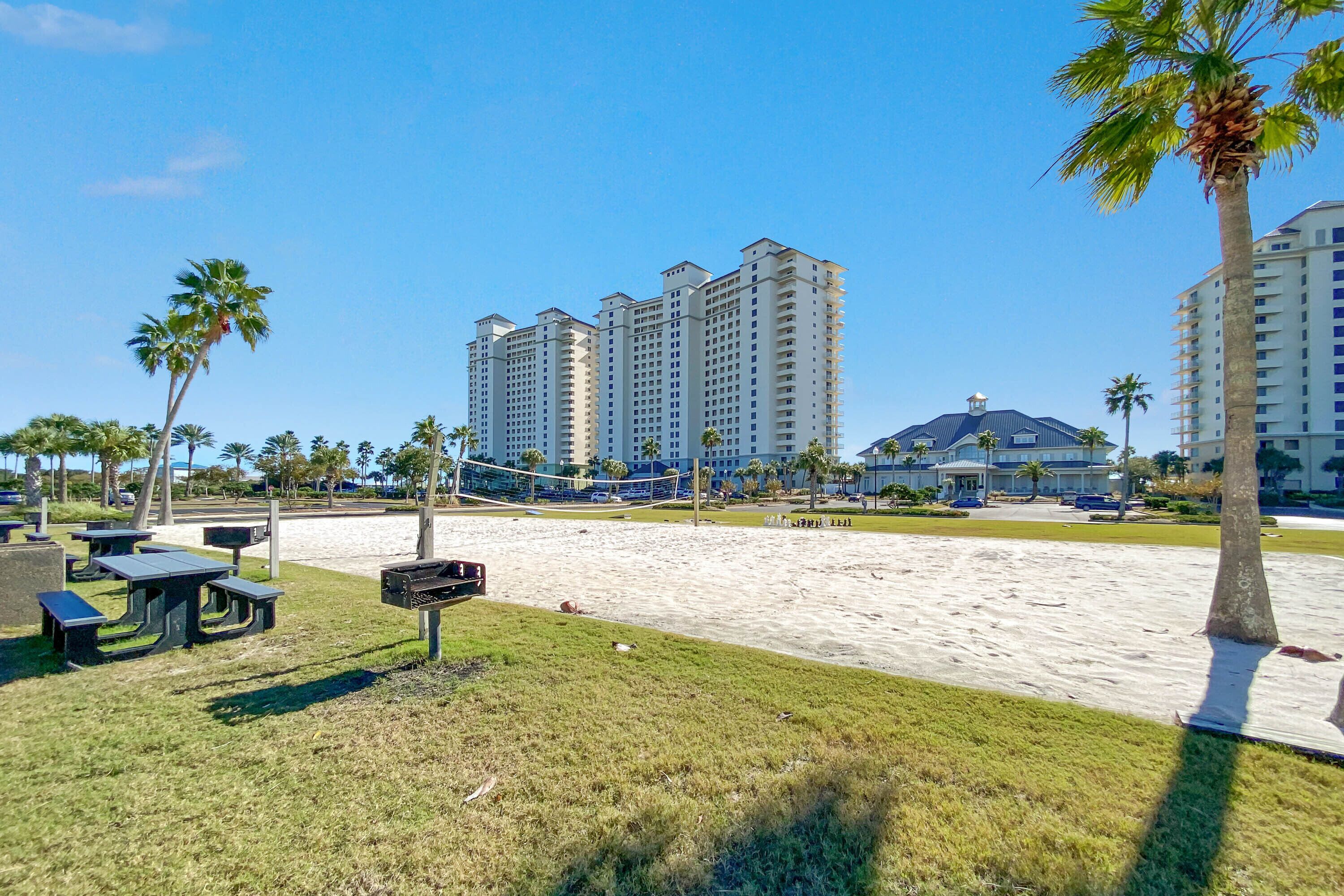 A beach area with picnic tables, a volleyball net, and tall buildings in the background.