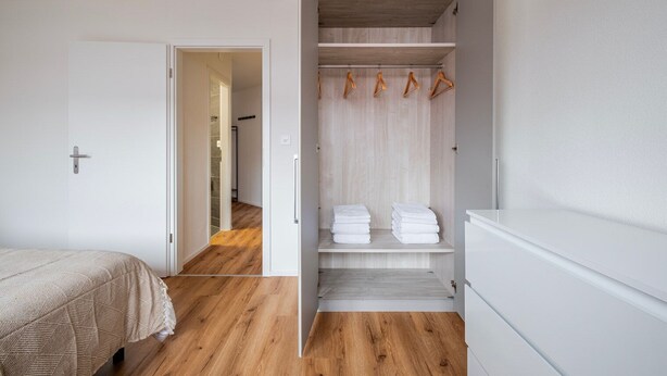 A bedroom with a bed, wooden flooring, a white wardrobe, and a hallway leading to another room. - Blueground | Dietikon, balcony, nr Lindenplatz (Zurich)
