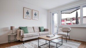 A modern living room with a sofa, a coffee table, a side table, and a chair. There are two framed artworks on the wall, and a window with a view of buildings outside. - Blueground | Dietikon, balcony, nr Lindenplatz (Zurich)