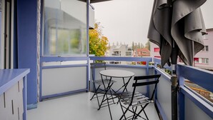 Un balcón con mesa y sillas, barandal azul y vistas a edificios y árboles.