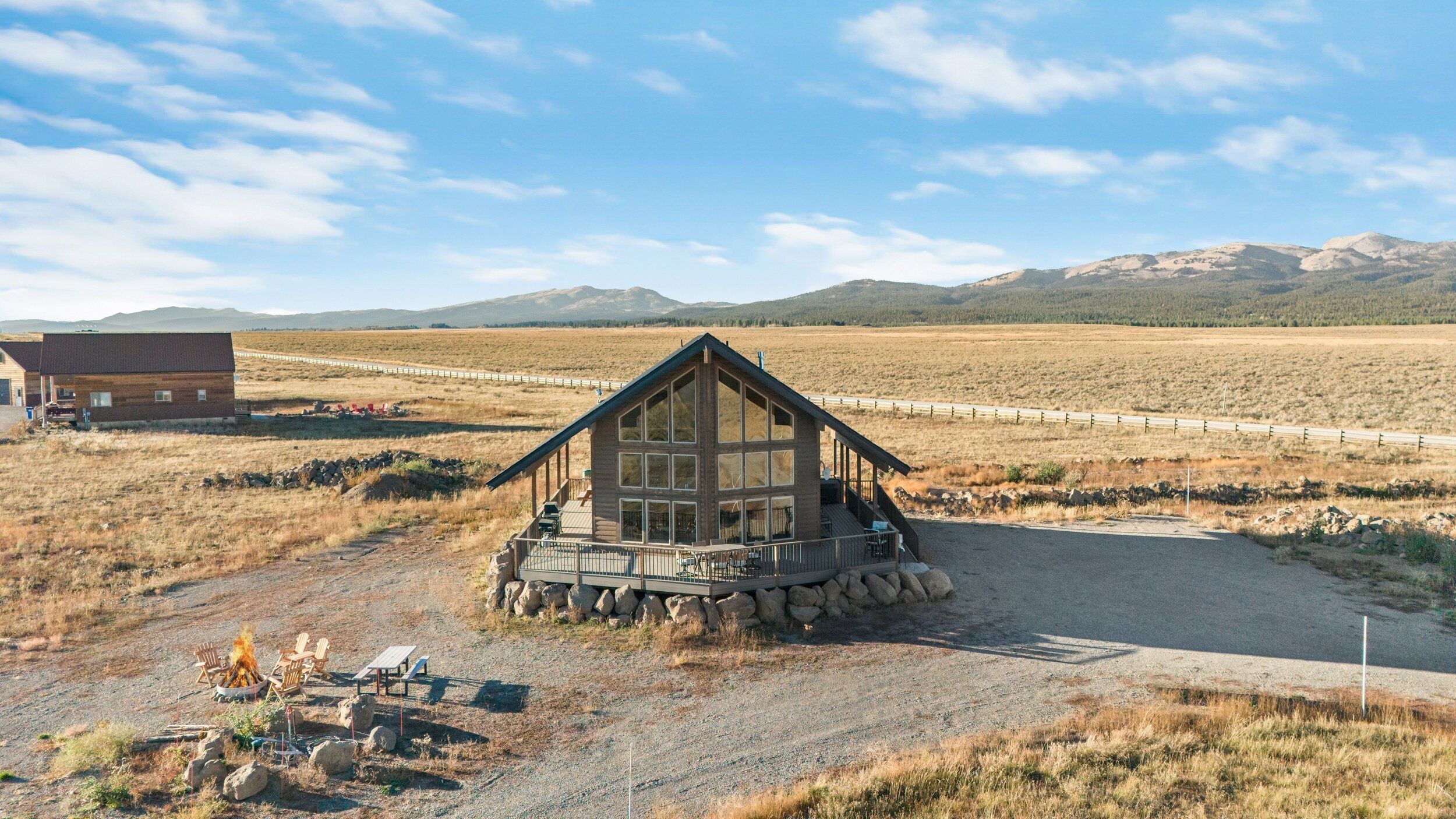 A lodge with a stone foundation and a wooden deck, surrounded by a dry grass field and mountains in the background.