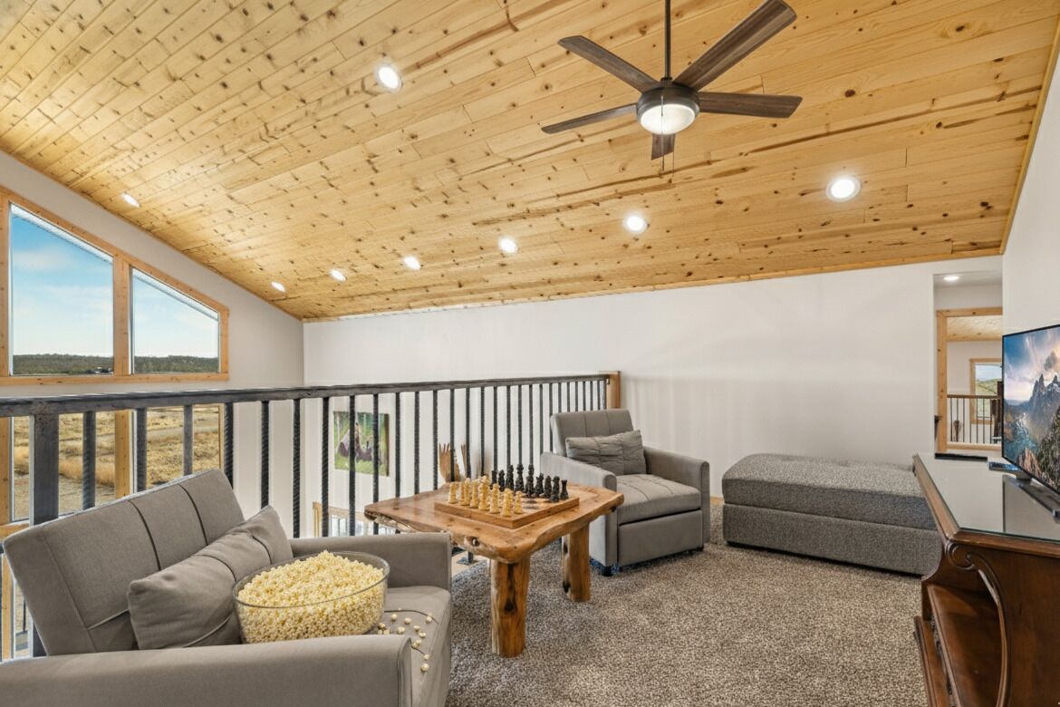A spacious room with a wooden ceiling, a large window, a balcony, a sofa, a coffee table, and a television.