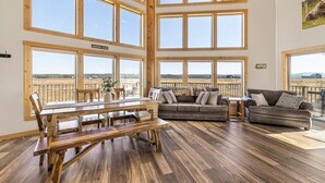 A spacious room with a large wooden dining table, a sofa, and a couch. There are multiple large windows providing a view of the outdoors. - Eagle Eye Retreat: AC, EV, Hot tub, Minutes to reservoir, 35 miles to W Yellowstone (Island Park)