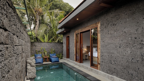 Un área de piscina con un muro de piedra, un rincón de descanso con cojines y una terraza de madera.