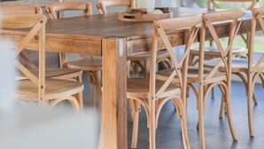 Une table à manger en bois, entourée de chaises en osier, dans une pièce lumineuse dotée de grandes fenêtres.