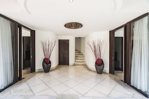 A spacious hallway with a tiled floor, two large potted plants, a wooden door, and a staircase leading to another level.