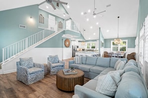 A spacious living room with a large L-shaped sectional sofa, a round coffee table, and two patterned armchairs. A staircase with a railing leads to an upper level.