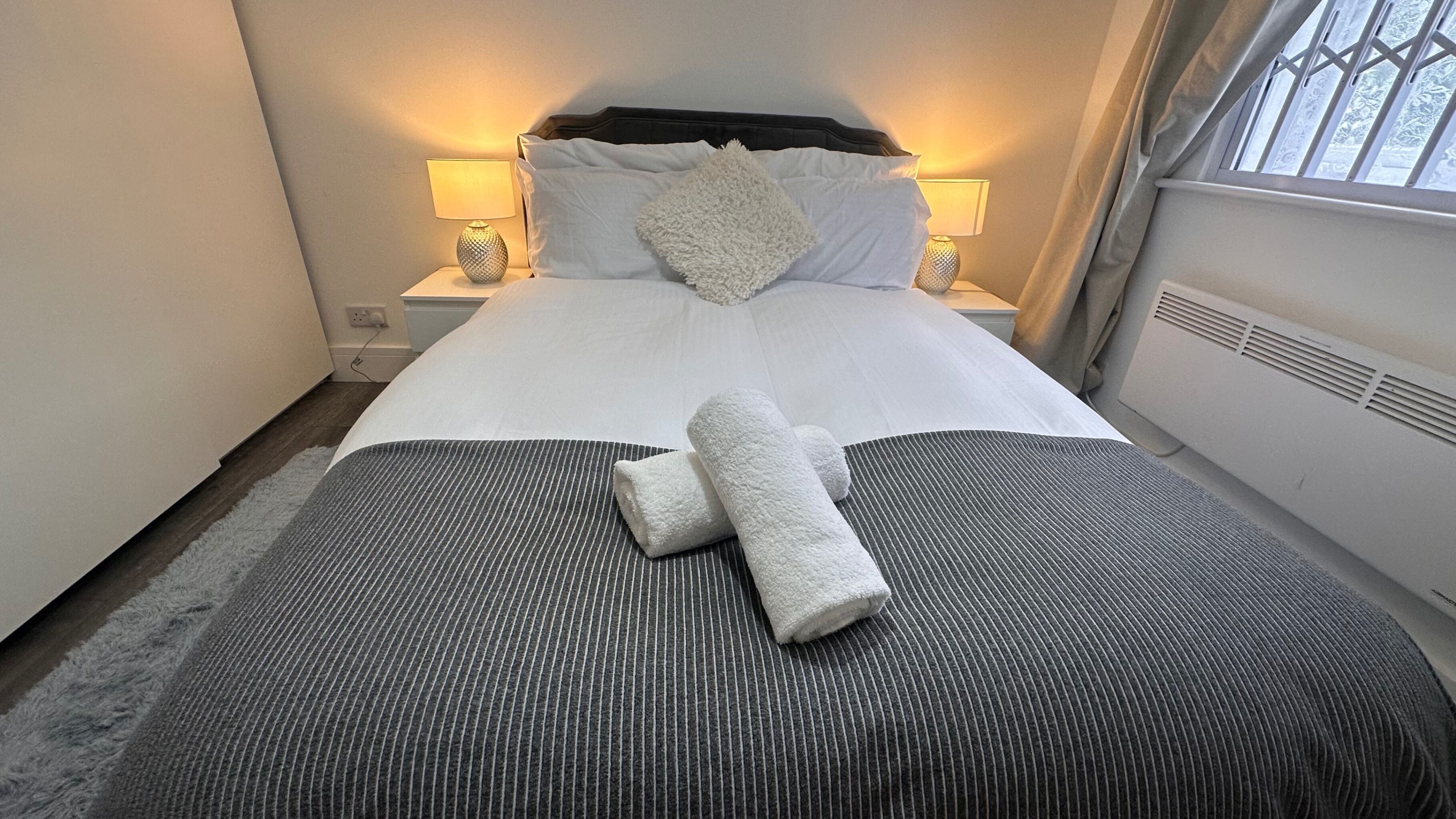 A neatly made bed with white linens, a grey striped blanket, and two rolled towels. There are two bedside lamps, a window with curtains, and a radiator.