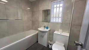 A modern bathroom with a bathtub, sink, toilet, and a window with a view. - Apartment - Sleeps 6 - City Base - District Line (Dagenham East)