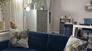 A modern living room with a blue sofa, two decorative pillows, a wooden floor, a radiator, a desk with a chair, and a shelf with books and decorative items. - Charming apartment in a great location. Near to the fun of Hartford (Hartford)