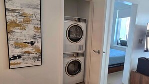 Une buanderie équipée de deux machines à laver à chargement frontal superposées, une porte donnant sur une chambre avec un lit et une fenêtre, et un téléviseur fixé sur un meuble en bois.