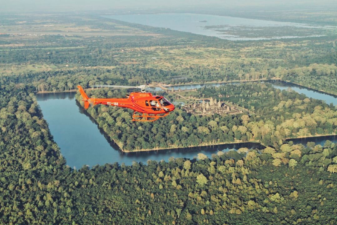 Um helicóptero sobre uma área arborizada, com um corpo d'água e uma construção.