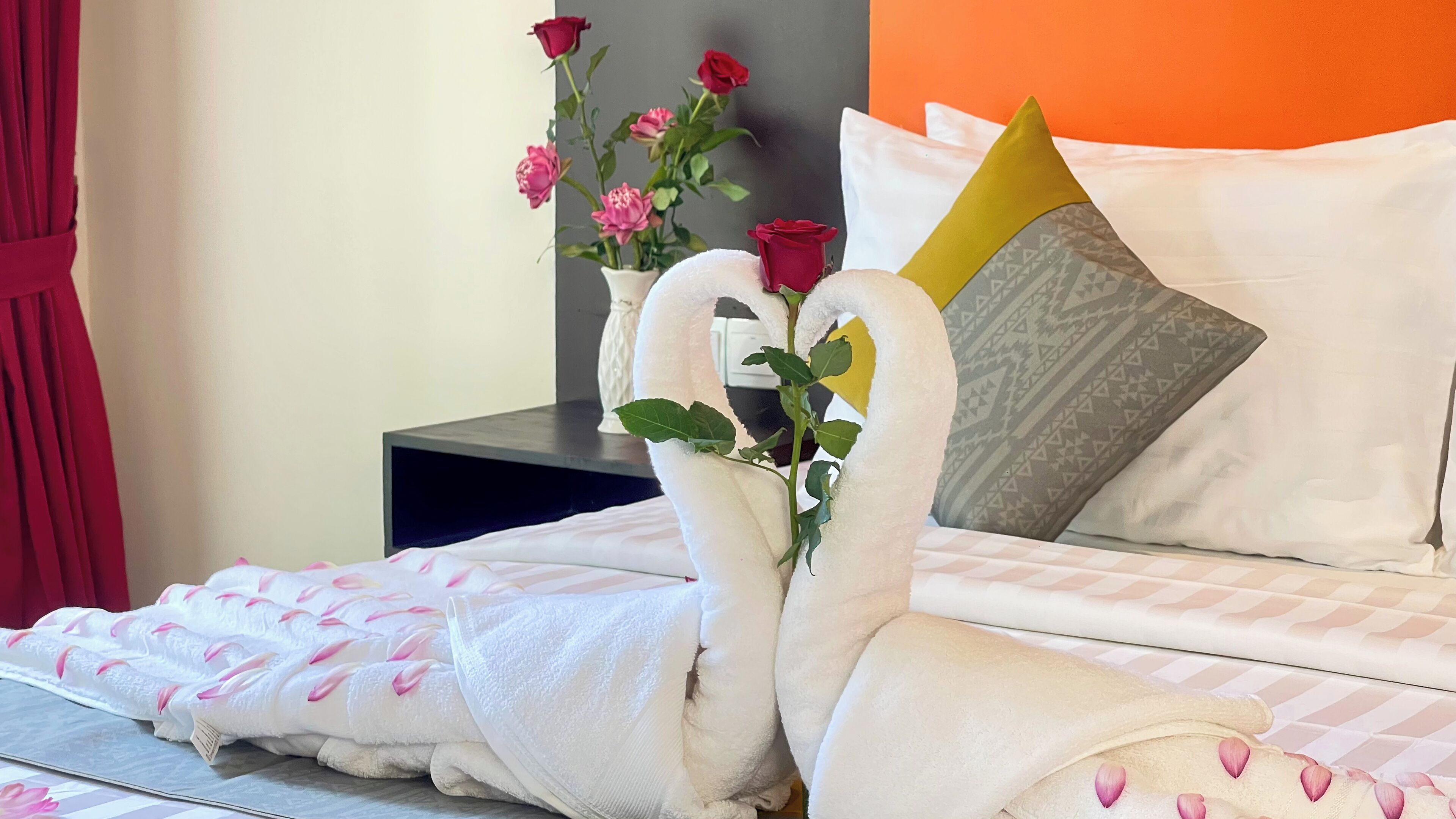 Un lit avec une literie blanche et des motifs floraux roses, une table de chevet avec un vase contenant des roses roses et rouges, et un coussin orné d’un motif géométrique.