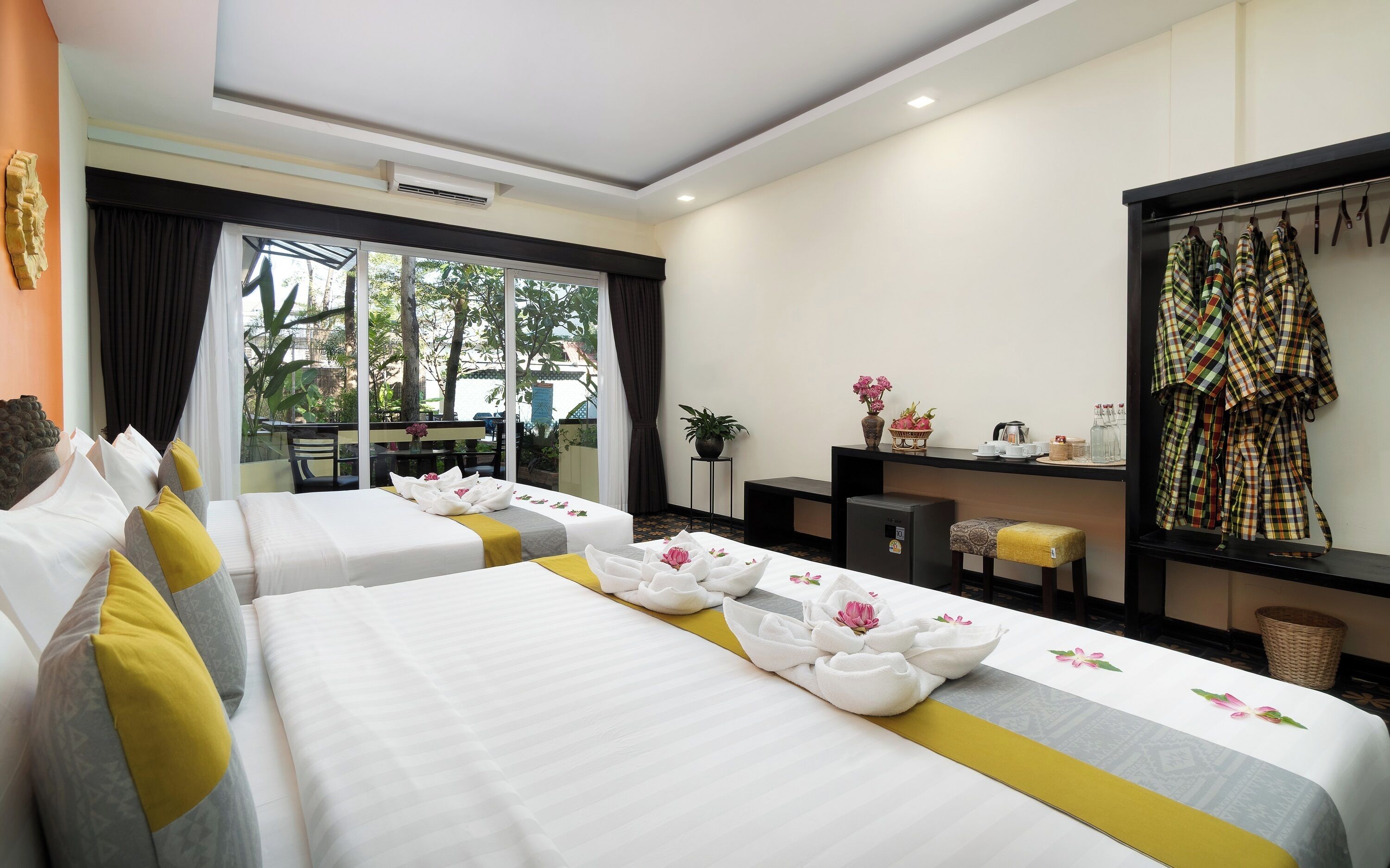 A hotel room with two beds, a desk, a chair, a small table, a mirror, and a window with a view of trees.