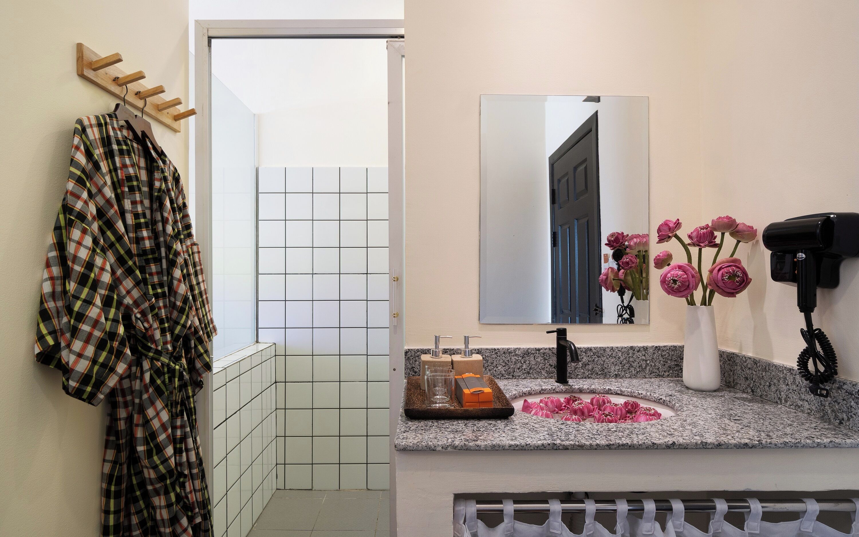 Une salle de bain avec un sol carrelé, un lavabo avec un miroir au-dessus, un sèche-cheveux fixé au mur et un porte-vêtements.