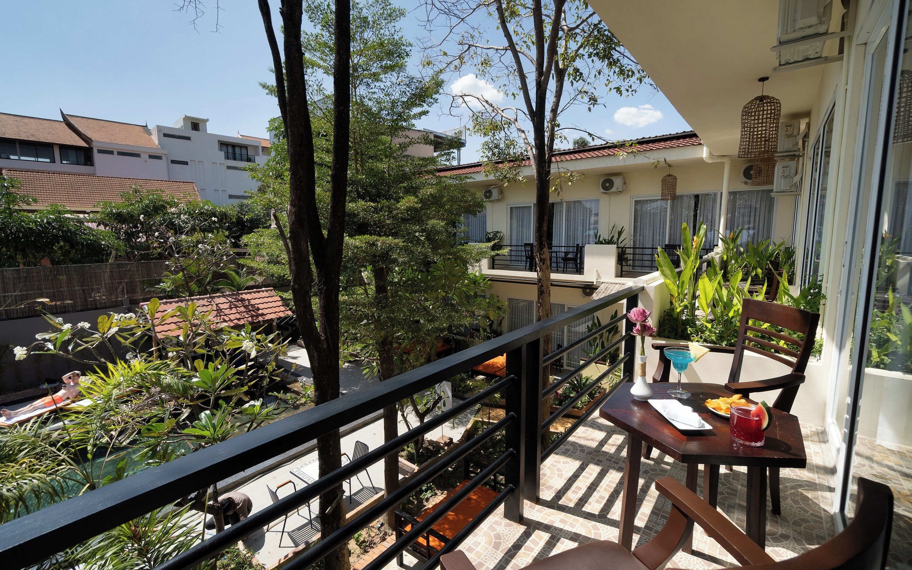 A balcony with a table, chairs, and potted plants. There is a view of a building with a tiled roof and a fence in the background.