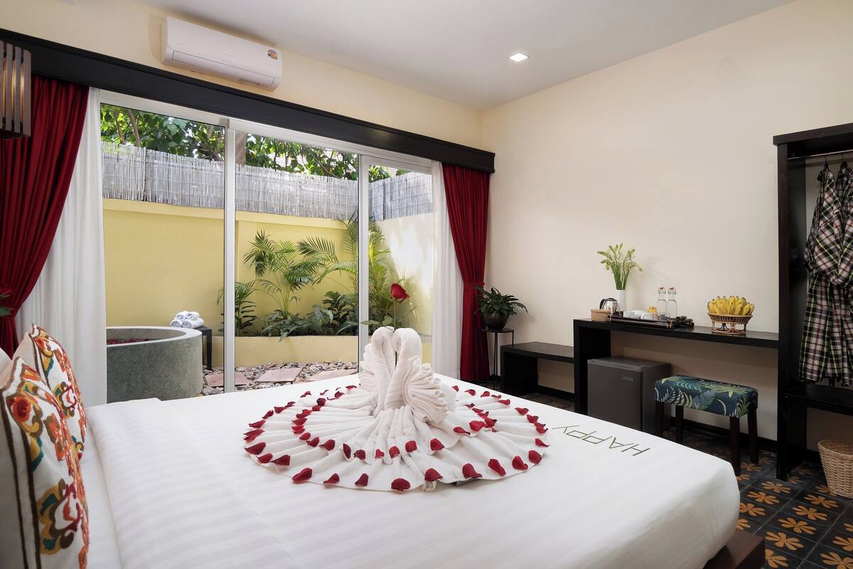 Une chambre d’hôtel avec un lit parsemé de pétales de rose, un bureau avec un ordinateur portable, une petite table sur laquelle est posé un vase de fleurs, et une vue sur la verdure à l’extérieur.