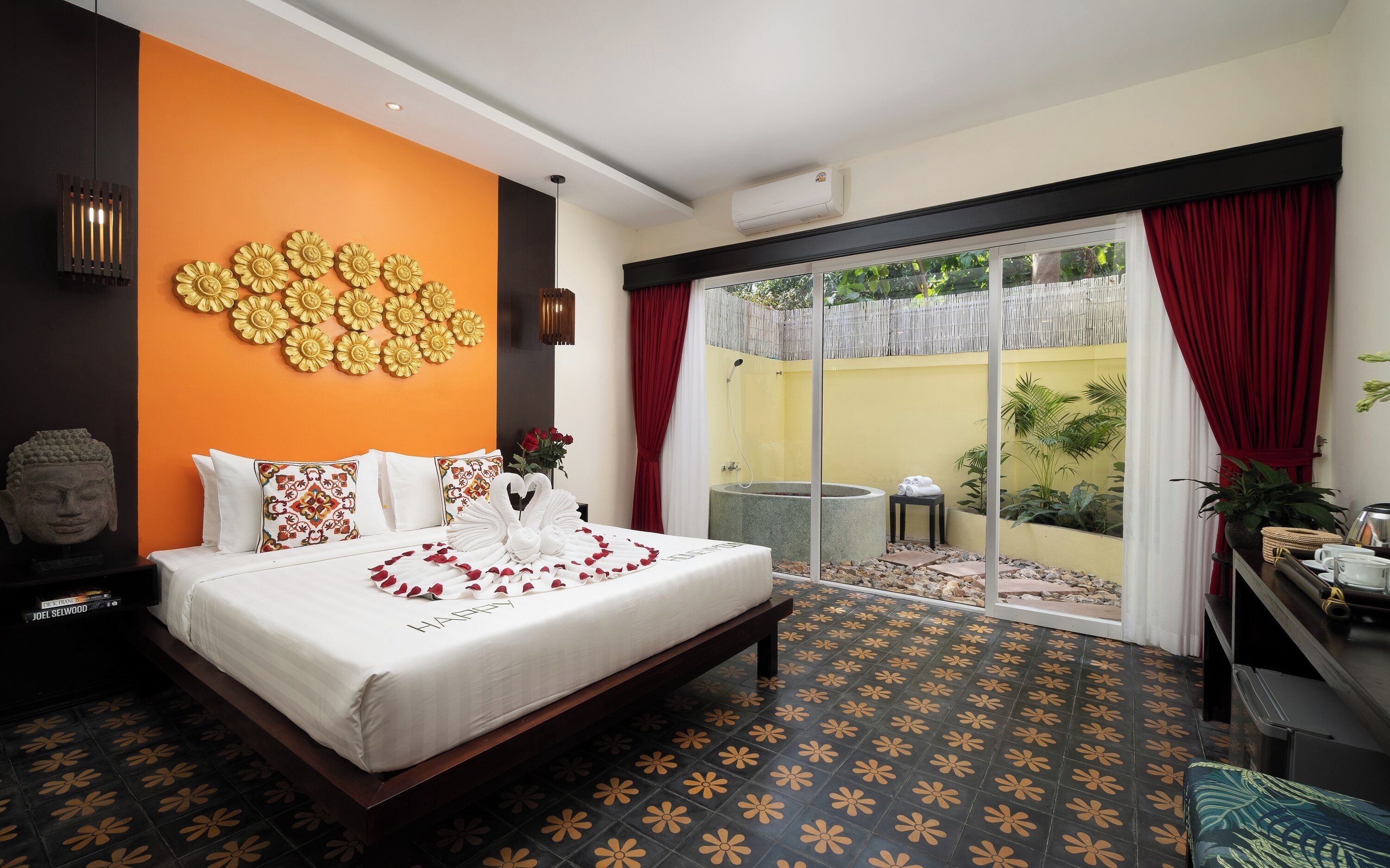 A hotel room with a bed, a Buddha statue, a wall with flower decorations, a shower area, and a balcony with plants.