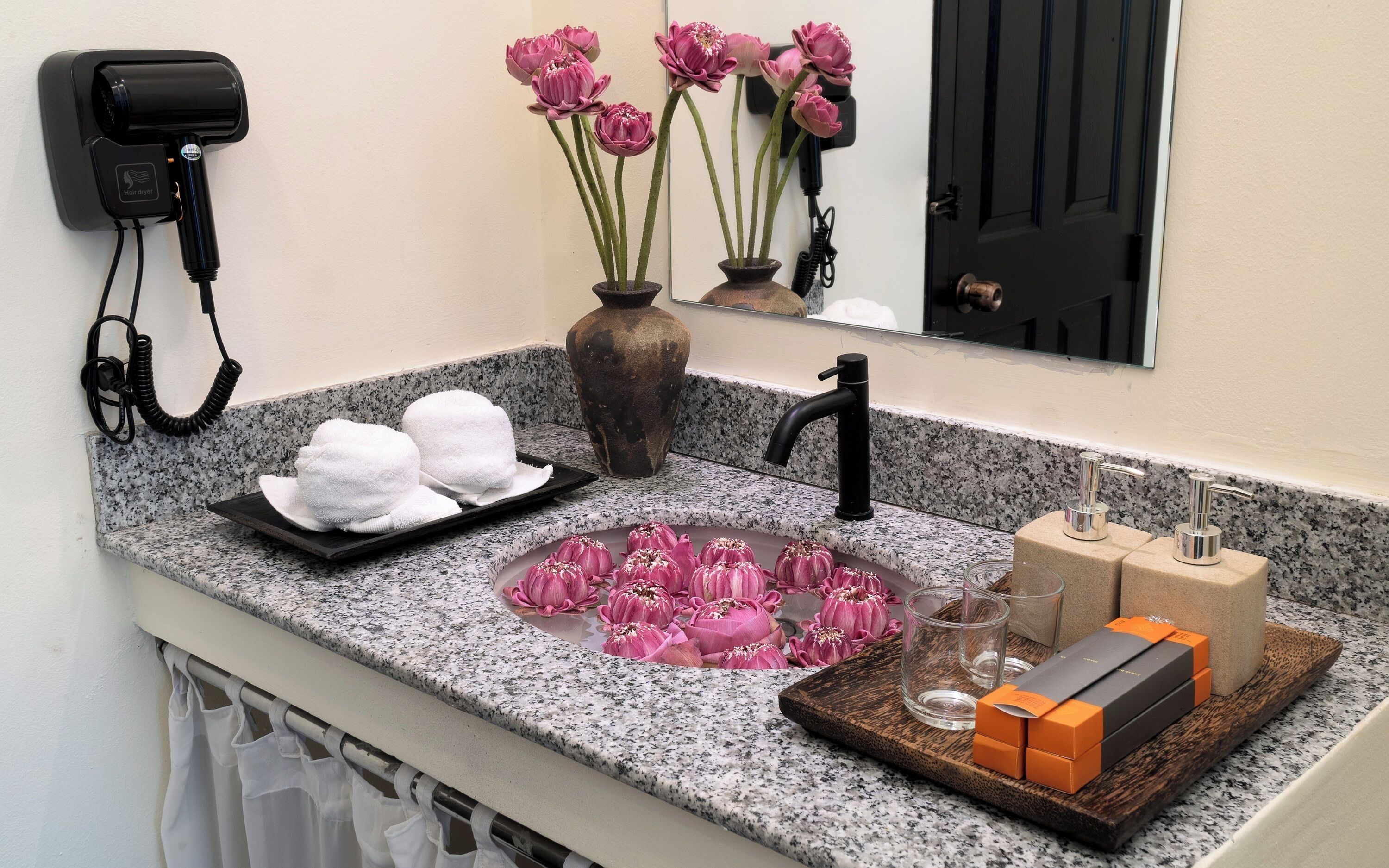 A bathroom with a granite countertop, a hairdryer, a vase with pink flowers, a sink, and toiletries.