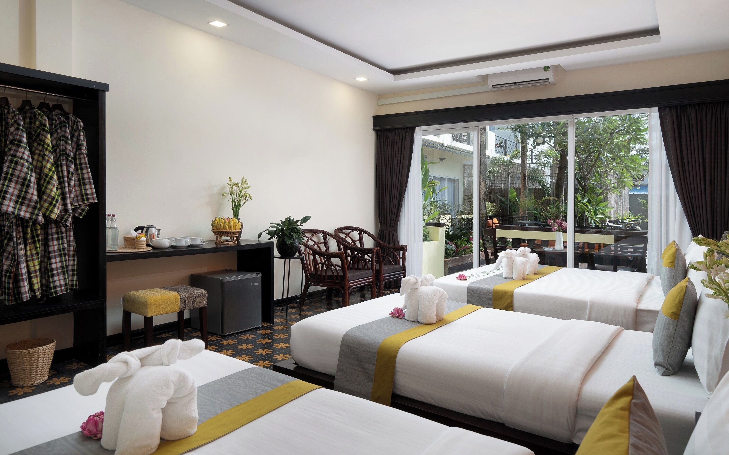 A hotel room with two beds, a desk with a computer, a chair, a small table with a plant, and a window with a view of greenery.
