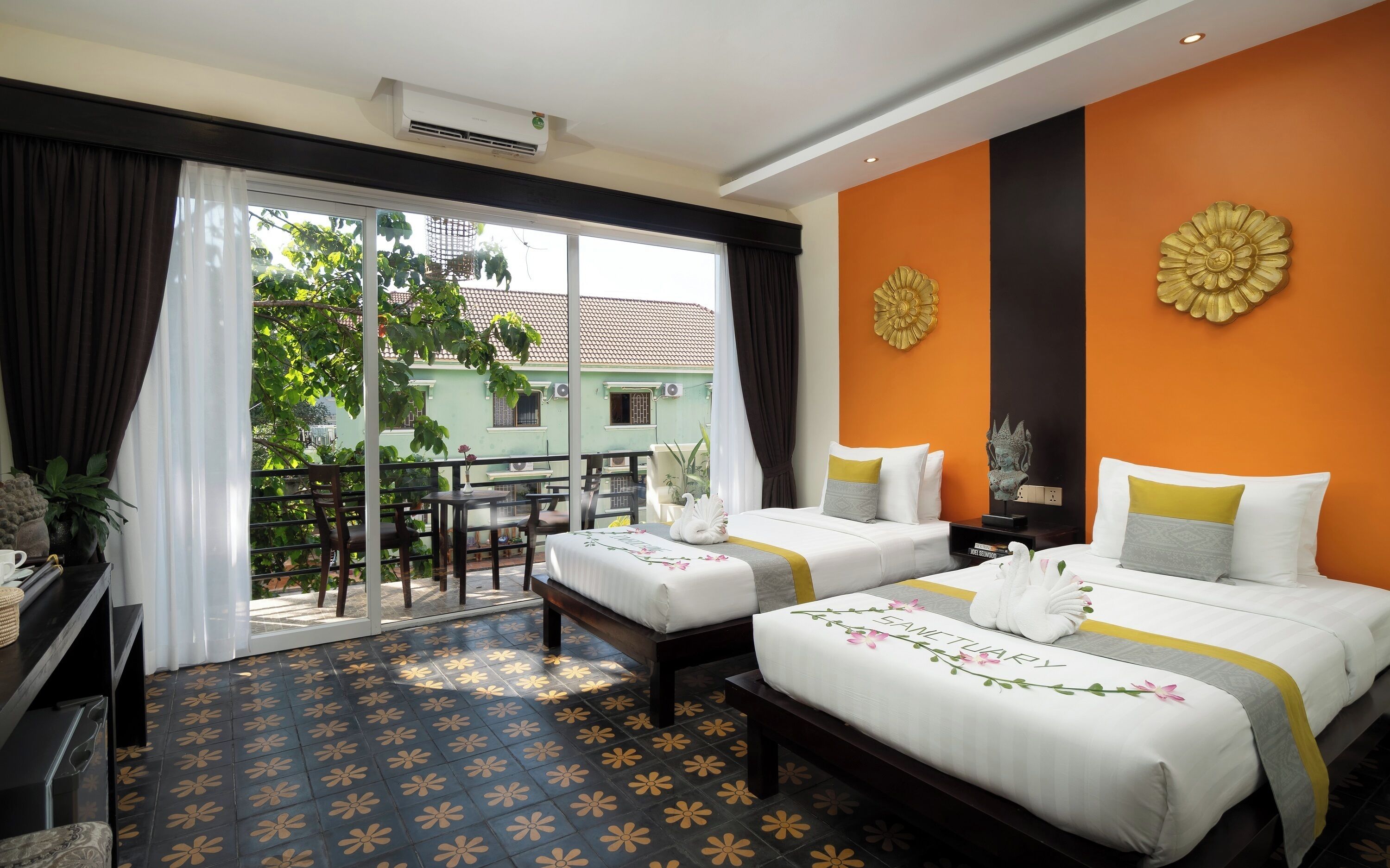 A hotel room with two beds, a balcony with a view of a green building, and a decorative wall piece.