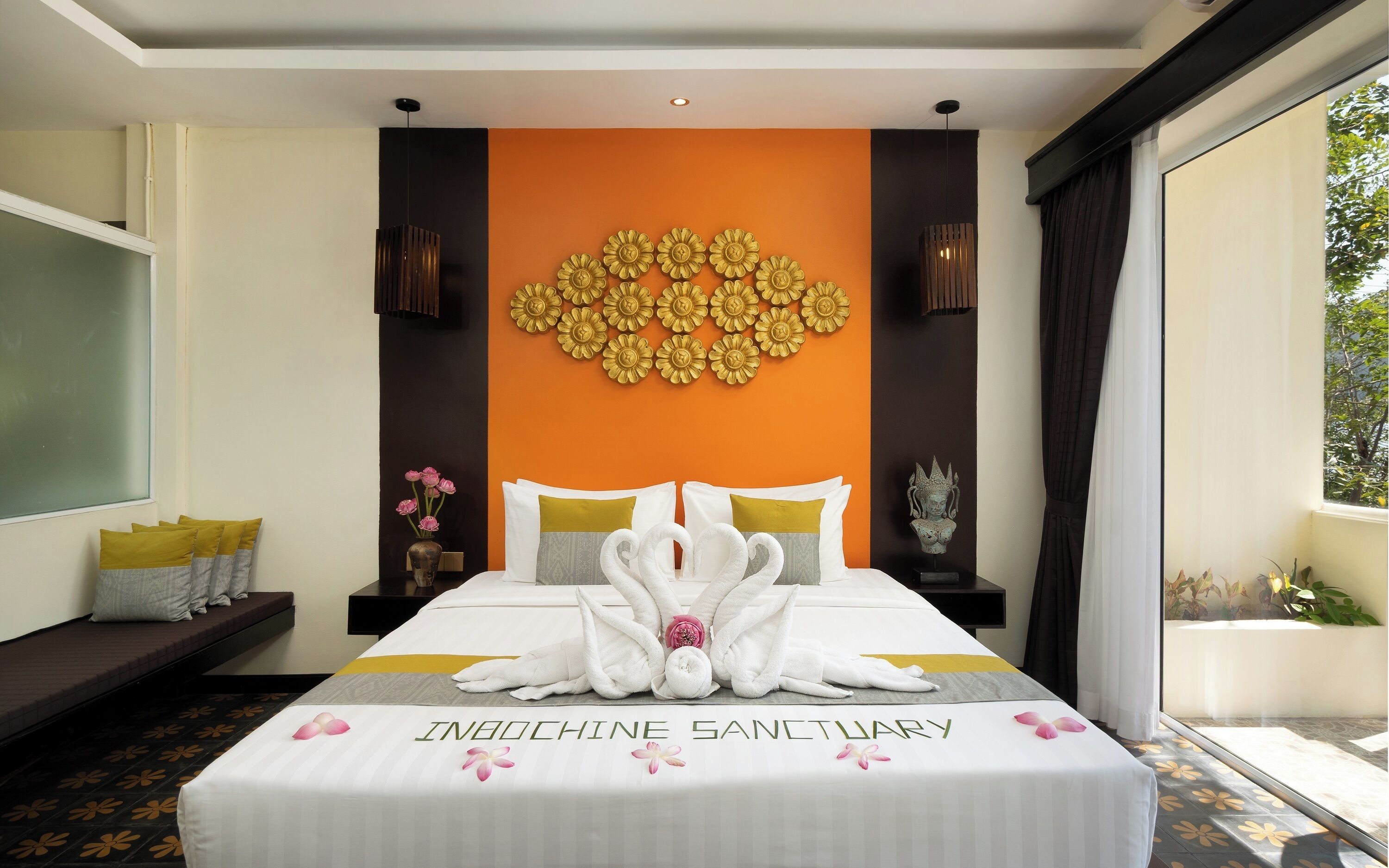 Quarto de hotel com cama, almofadas, um vaso com flores, um objeto decorativo na parede e uma janela com cortinas.