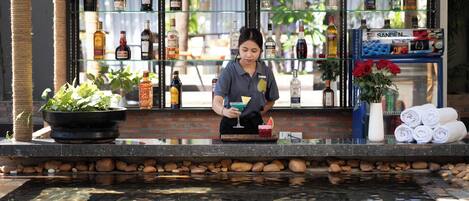 Uma área de bar com um bartender, diversos frascos de bebidas alcoólicas, uma planta em vaso e um pequeno elemento decorativo com água.