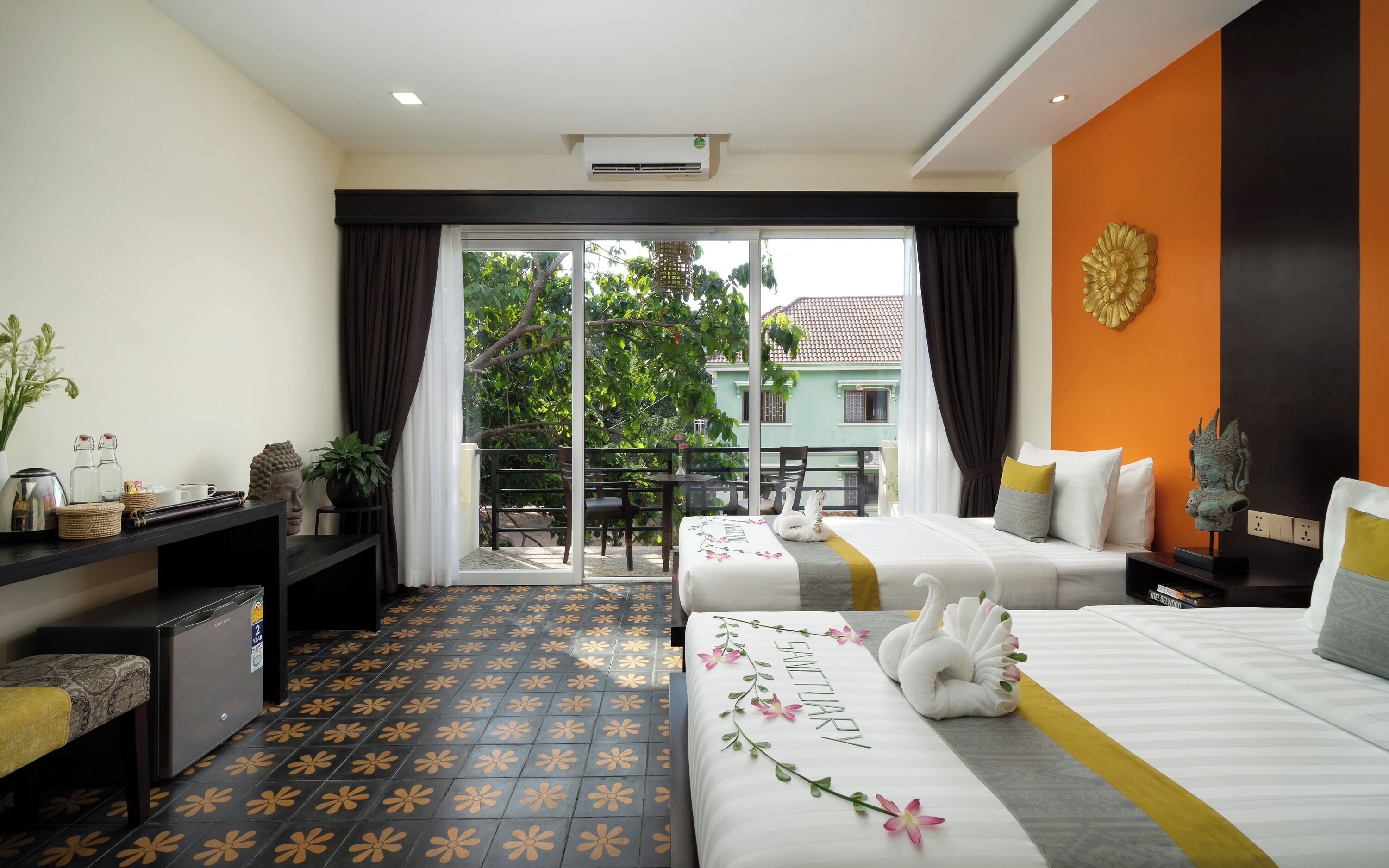 A hotel room with two beds, a desk, a small table, and a balcony with a view of a building and trees.
