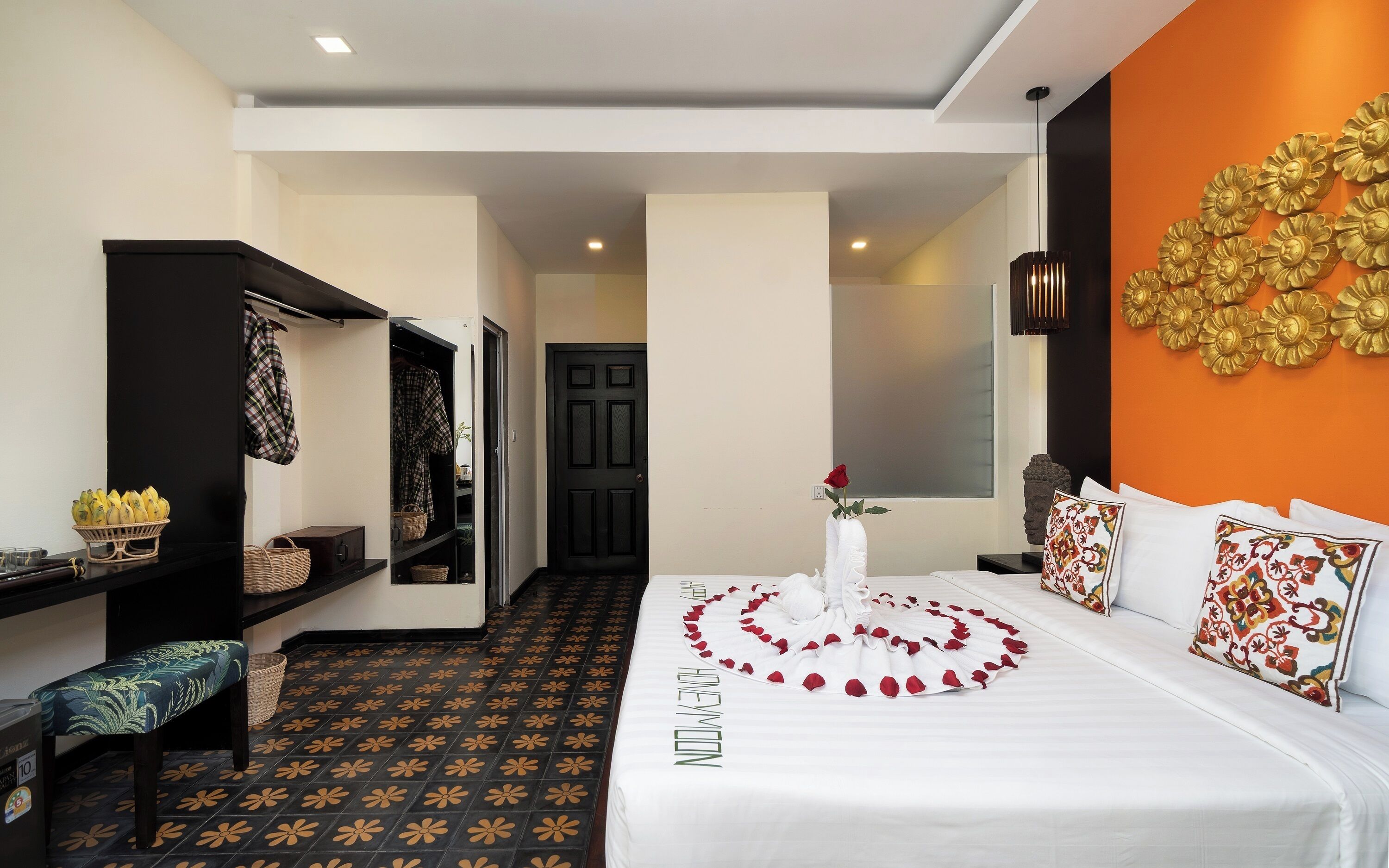 A hotel room with a bed adorned with rose petals and a single rose, a bench with a floral cushion, a wardrobe, and a wall with a decorative golden floral arrangement.