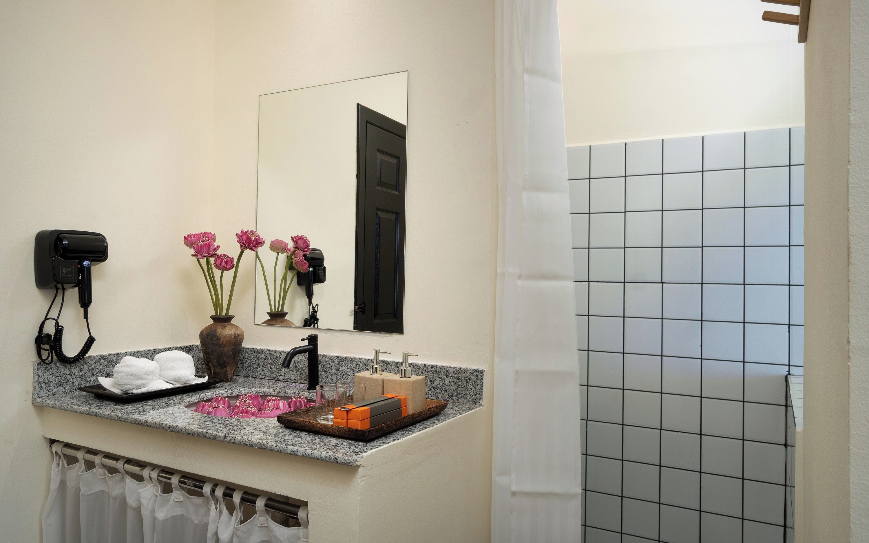 A bathroom with a vanity, a hairdryer, a mirror, a sink, and a shower area.